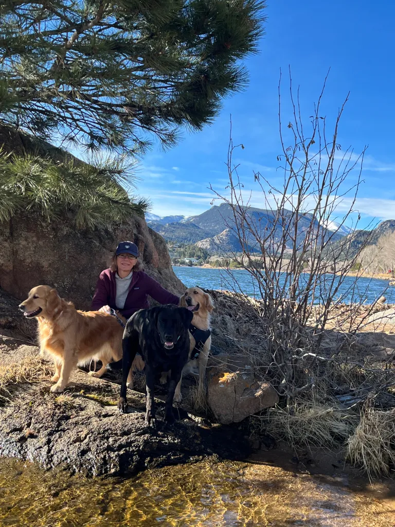 A woman with three dogs next to a lake with mountains in the background.