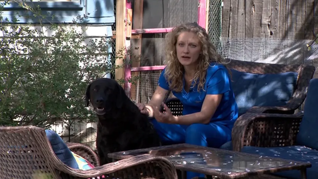 A woman in blue scrubs pets a black dog sitting on a patio.