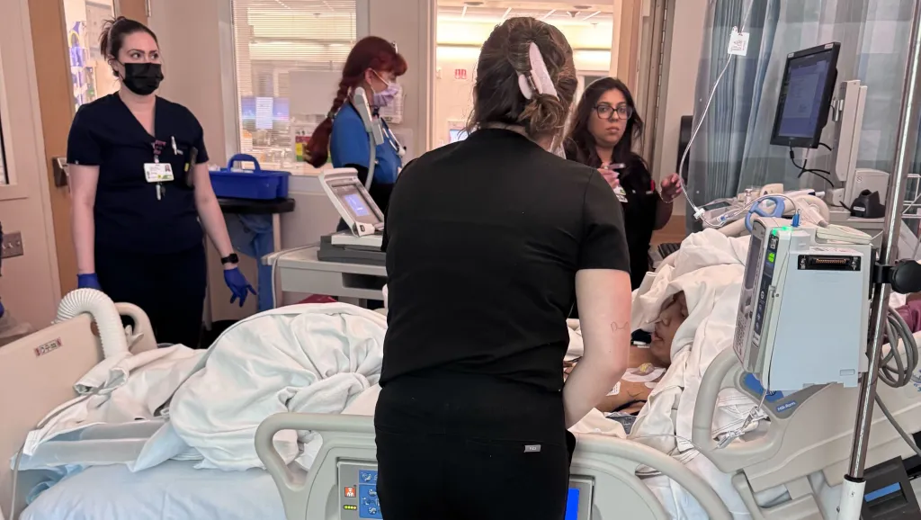 Comedian Alex Duong in a hospital bed surrounded by medical staff and equipment.