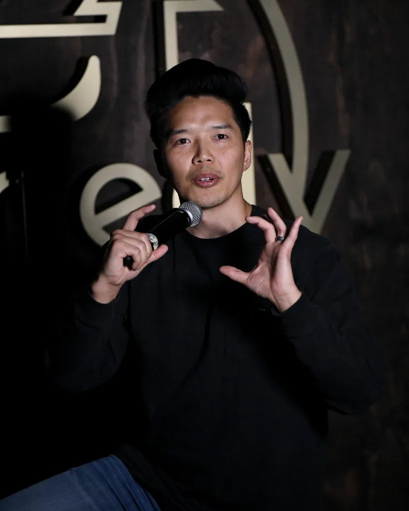 Comedian Alex Duong performing with a microphone in his left hand, making a gesturing motion with his right hand.