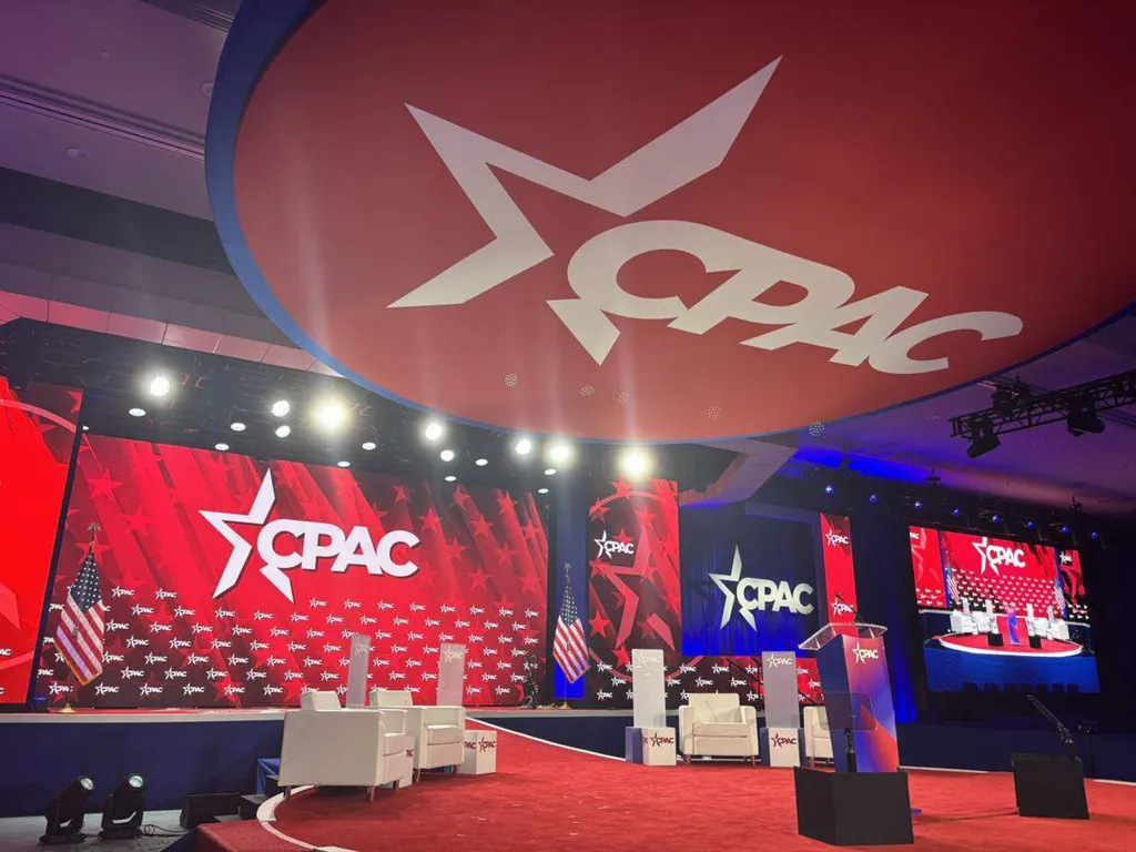 The CPAC conference stage with a large red backdrop featuring the CPAC logo, two American flags, and several white armchairs on a red carpet.