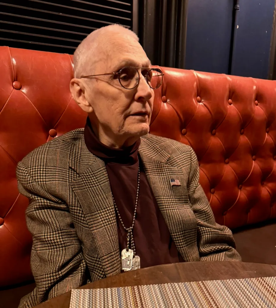 Richard Williams, 83, wearing glasses, a plaid jacket, and a maroon turtleneck with dog tags, sitting on a red tufted bench.