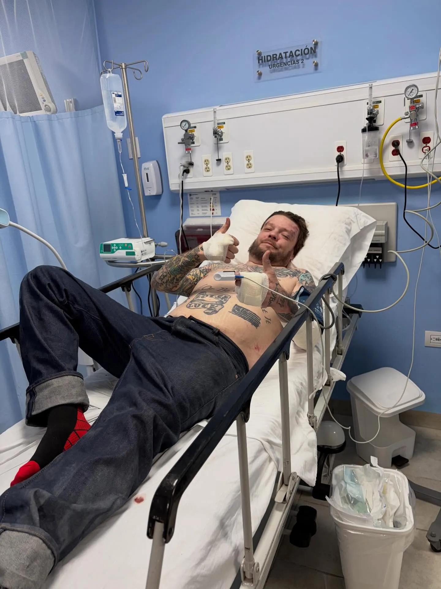 Corey Harrison in a hospital bed with an IV drip, giving a thumbs up.