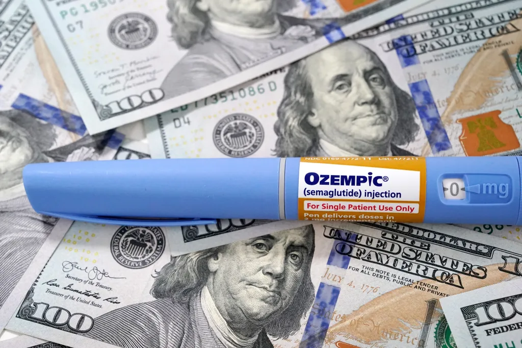 An Ozempic injection pen lies on a scattered background of one hundred dollar bills.