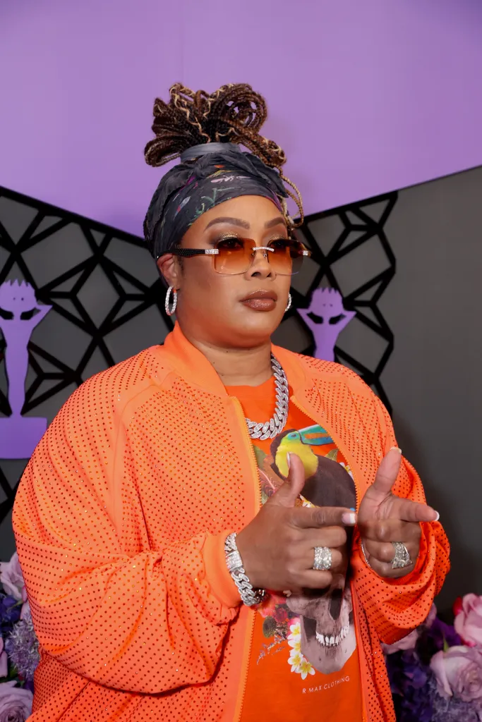 Da Brat in an orange sequined jacket over a t-shirt, with a patterned headscarf, sunglasses, and multiple rings.