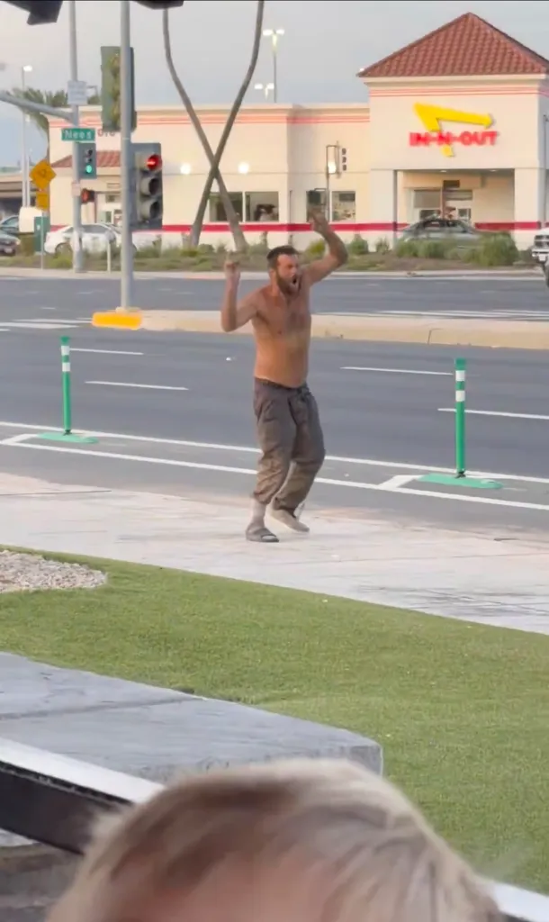 A shirtless man with his arms raised, screaming, on a sidewalk near a street with an In-N-Out Burger in the background.