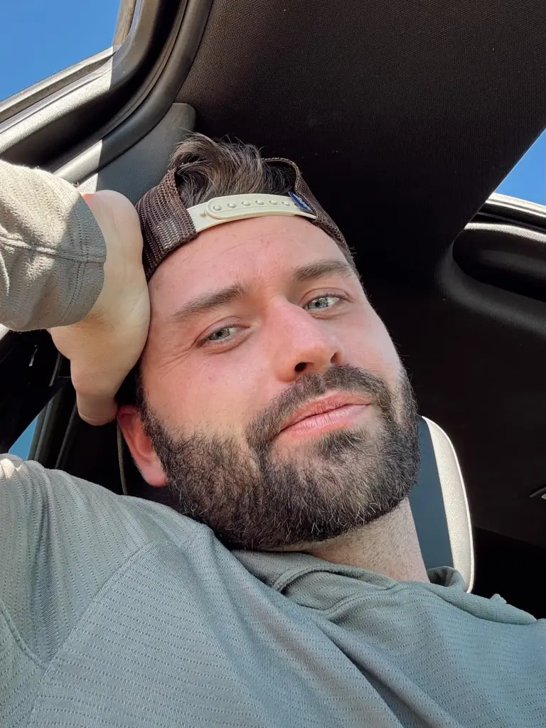 A man with a beard and baseball cap gazes at the viewer from inside a car.