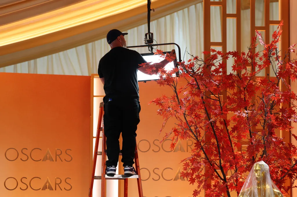 A worker on a ladder adjusts a light next to a decorative replica Oscar statue covered in plastic sheeting and a display of red maple leaves.