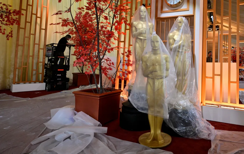 Oscar statues covered in plastic sheeting on a red carpet as a worker prepares the event space.