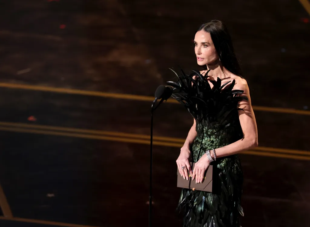 Demi Moore speaking onstage at the 98th Oscars, wearing a strapless green feather dress and holding a brown envelope.