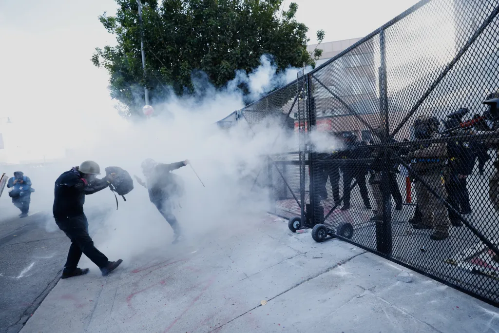 Demonstrators running through tear gas with a fence on the right.