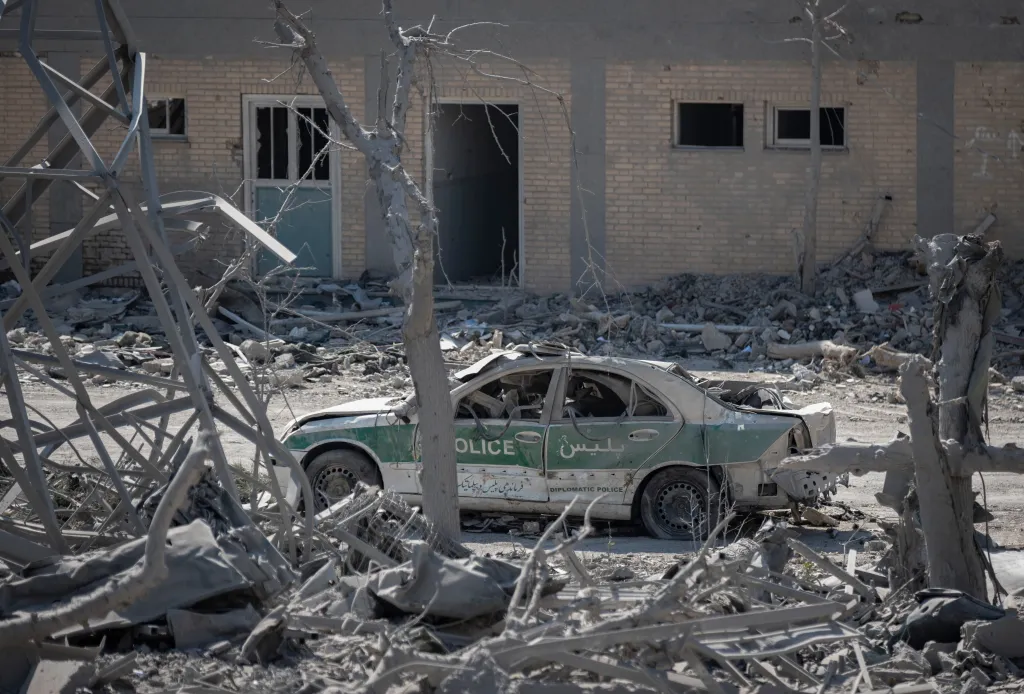 A destroyed police car sits amidst rubble in front of a damaged brick building.