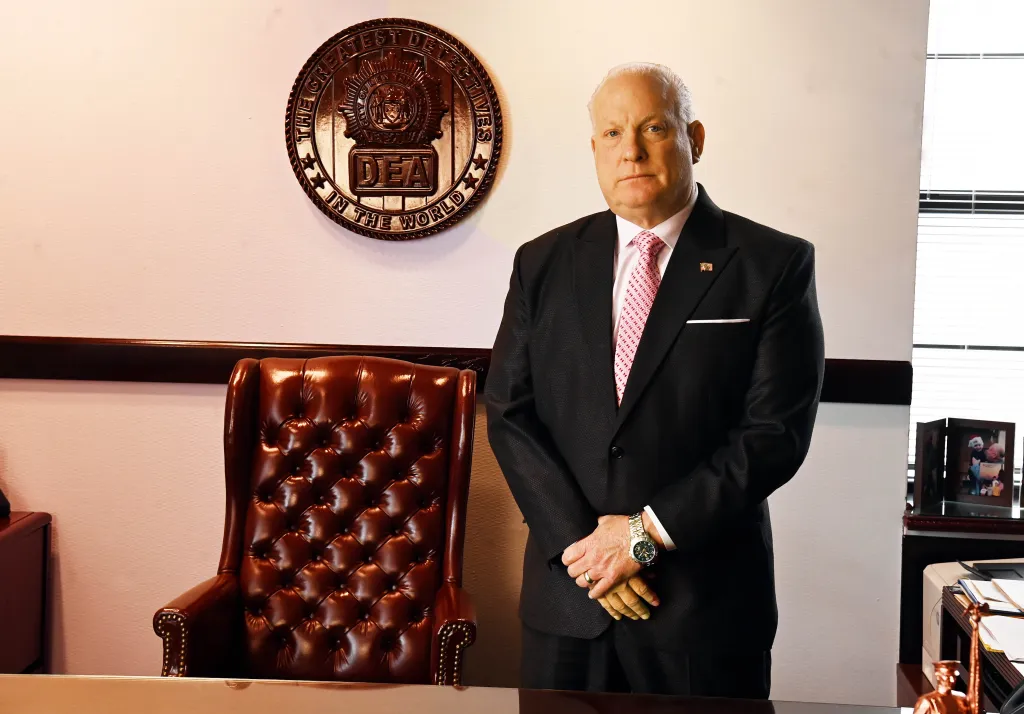 Detective Scott Munro, president of the Detectives' Endowment Association, stands in his office in front of a circular DEA emblem.