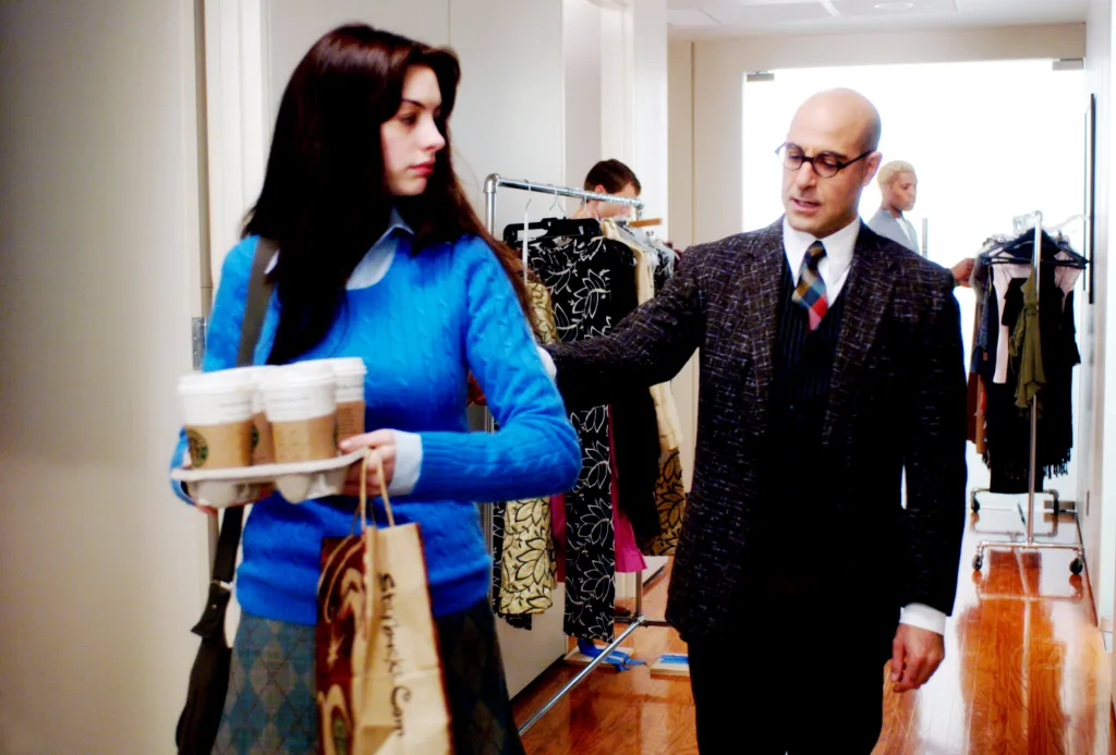 Anne Hathaway as Andy Sachs, in a blue sweater and grey skirt, carrying Starbucks coffees and Stanley Tucci as Nigel Kipling , in a black suit and red checked tie, in 