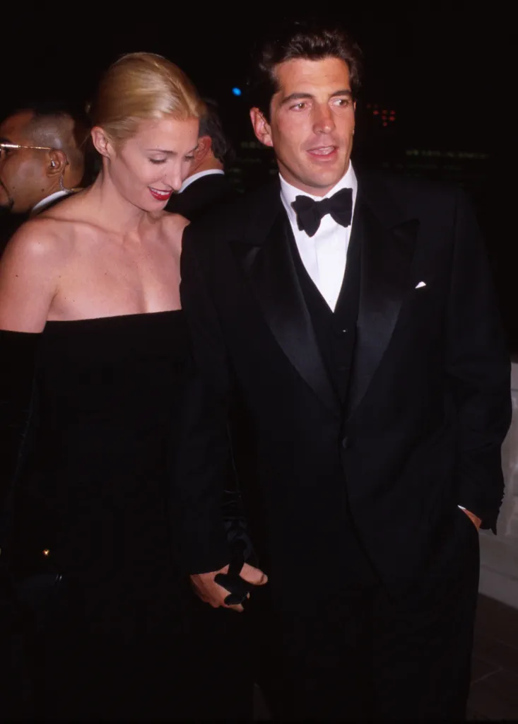 John F. Kennedy Jr. in a tuxedo and Carolyn Bessette in a black dress, holding hands at a formal event.