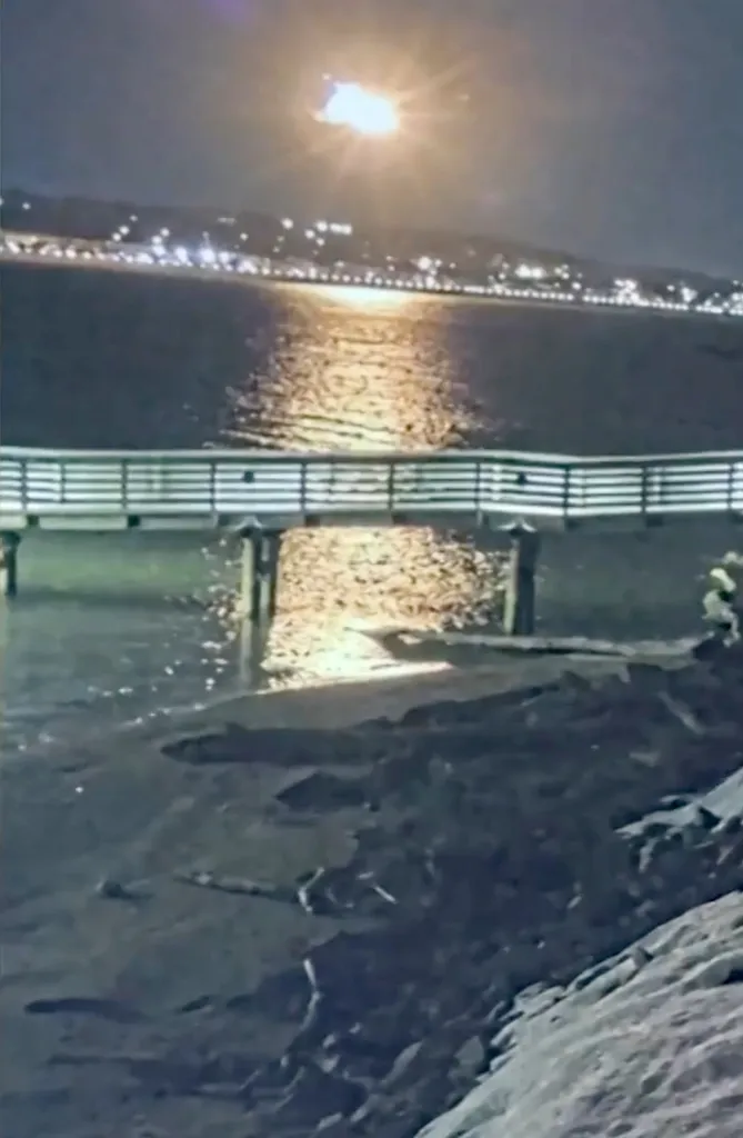 A fiery explosion illuminates the night sky over a body of water with a pier in the foreground.