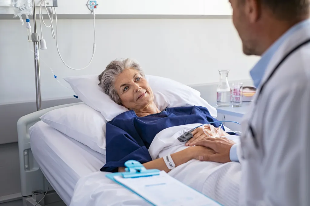 A doctor in a white coat holds the hand of a smiling senior woman in a hospital bed.