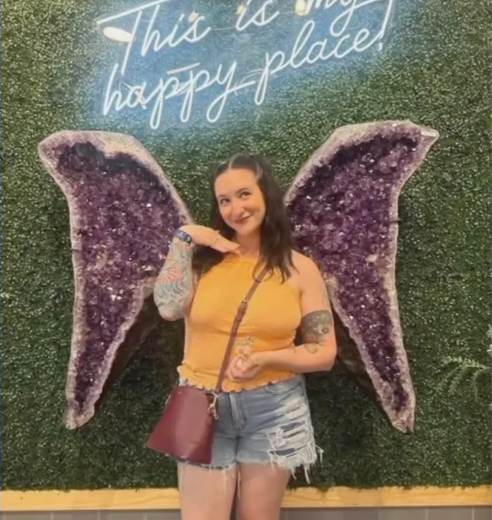 Rachel Long standing in front of amethyst wings, with a neon sign above that reads, 