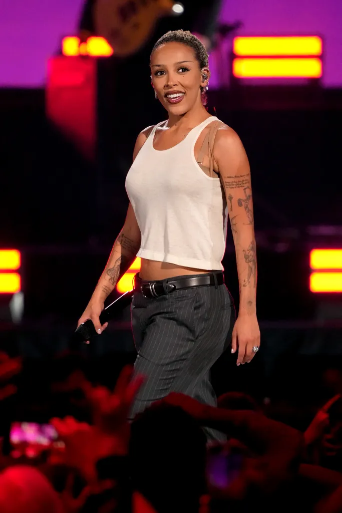 Doja Cat performing onstage, wearing a white tank top and pinstriped trousers, holding a microphone.
