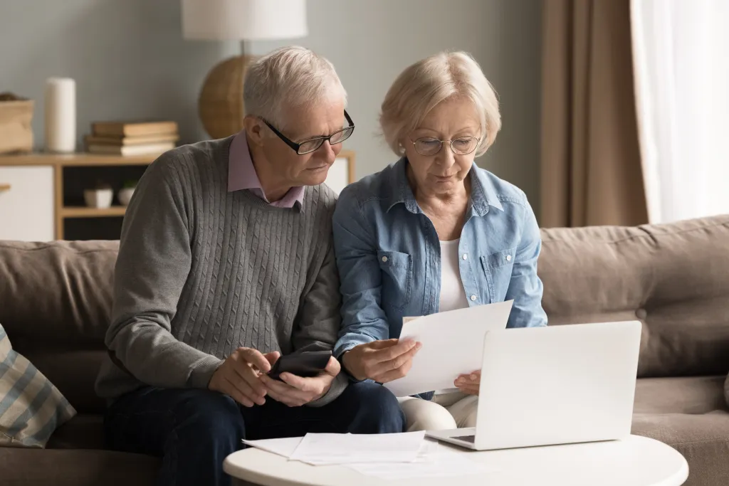 Elderly couple at home managing their finances using a laptop, calculator, and paperwork.
