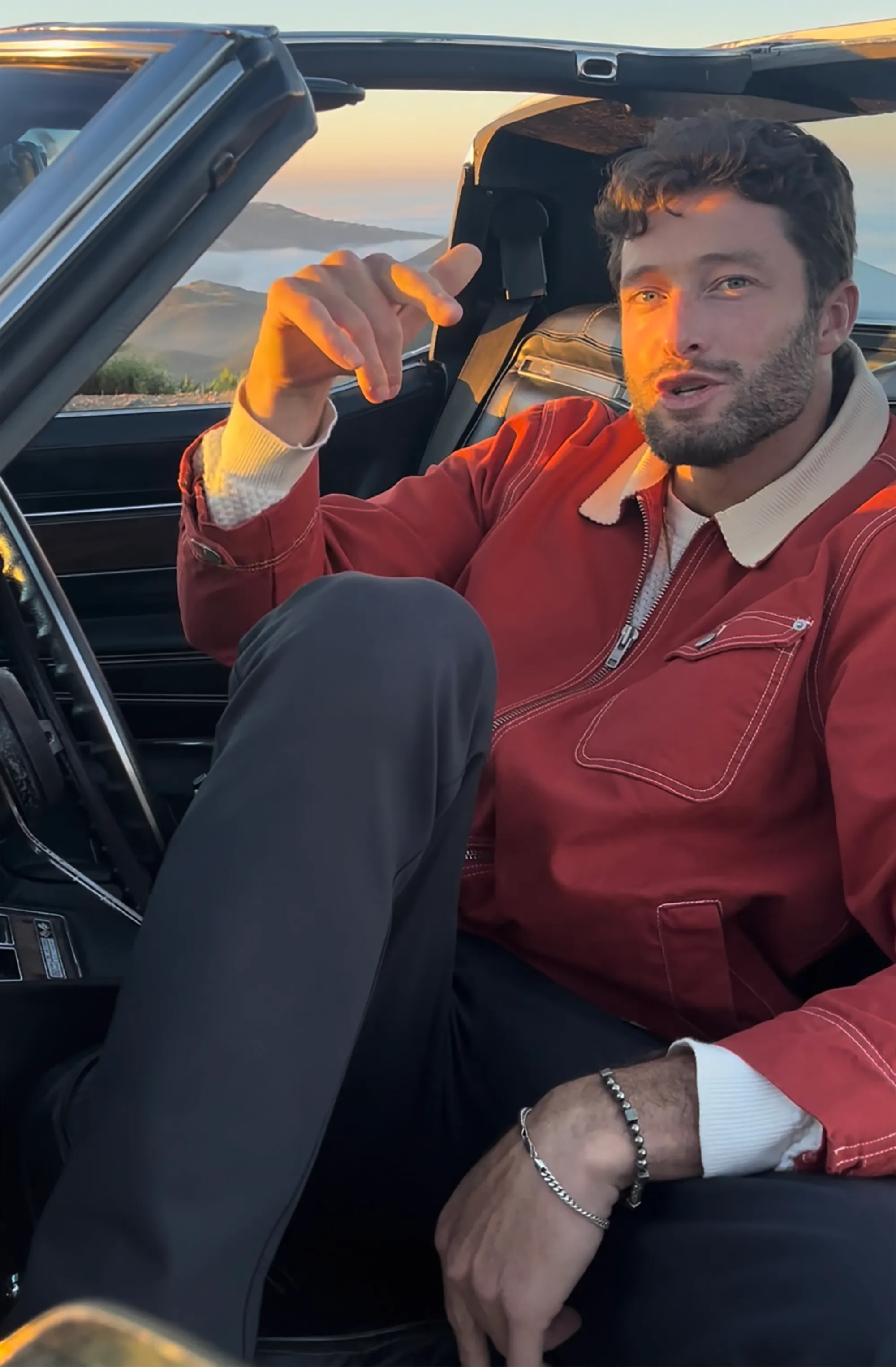 Doug Mason in a red jacket, seated in a car, singing along to his new song with mountains in the background.