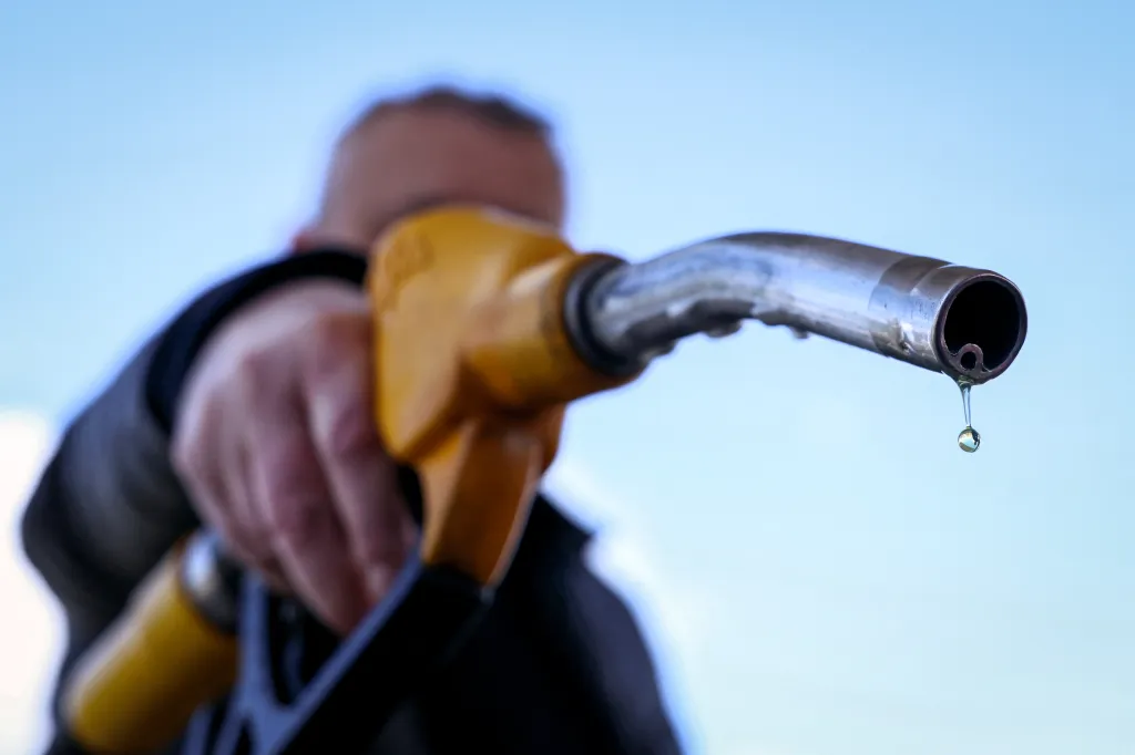 A person's hand holds a yellow and silver petrol pump nozzle with a single drop of petrol dripping from the end against a clear blue sky.