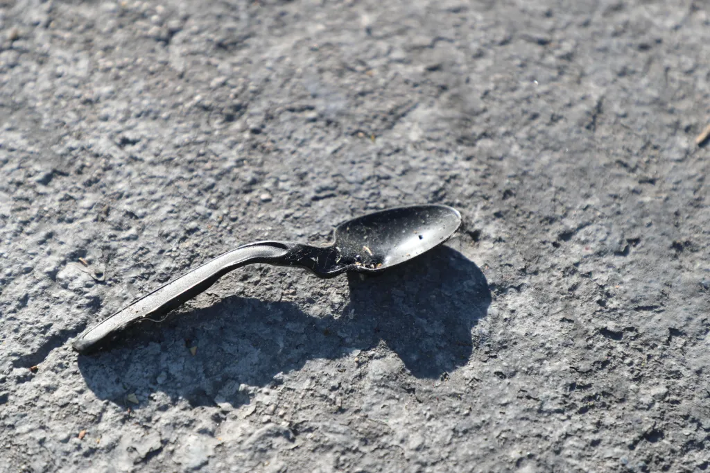 A black plastic spoon with a burned bowl lies on rough, grey ground.