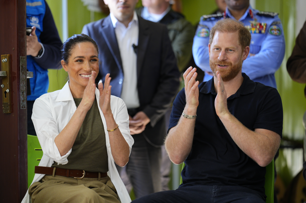 Meghan Markle, in a white shirt, brown t-shirt and khaki pants, and Prince Harry, in a black t-shirt and black pants, clapping during a music performance at the Za'atari refugee camp in Jordan.