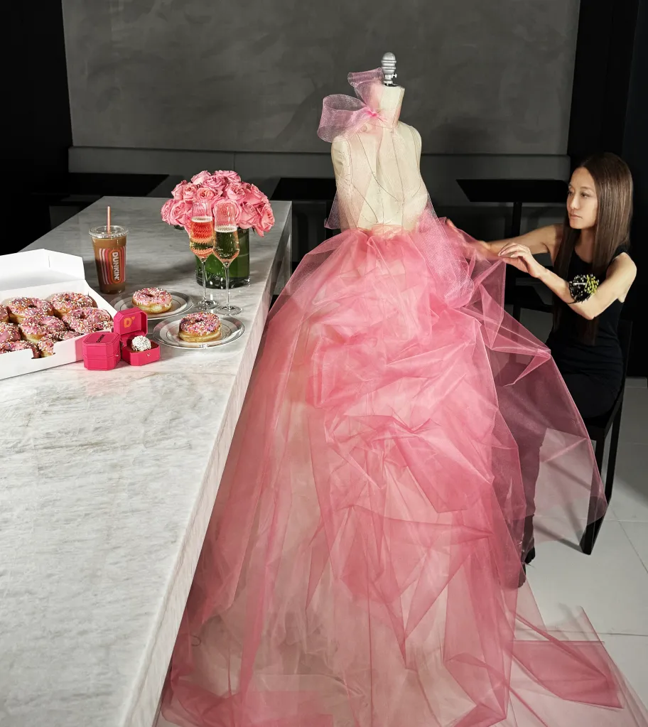 A woman adjusting a voluminous pink dress on a mannequin next to a table with Dunkin' donuts, drinks, and pink ring boxes.