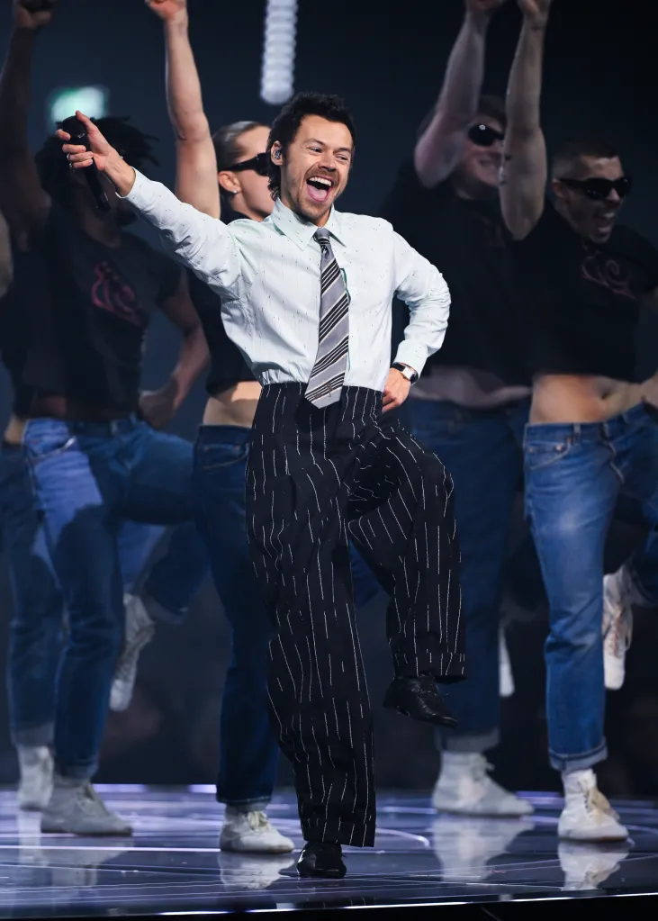 Harry Styles performing at The BRIT Awards, holding a microphone with his right hand raised, smiling and singing, wearing a light blue striped shirt, a striped tie, and black pinstriped trousers, with backup dancers behind him.