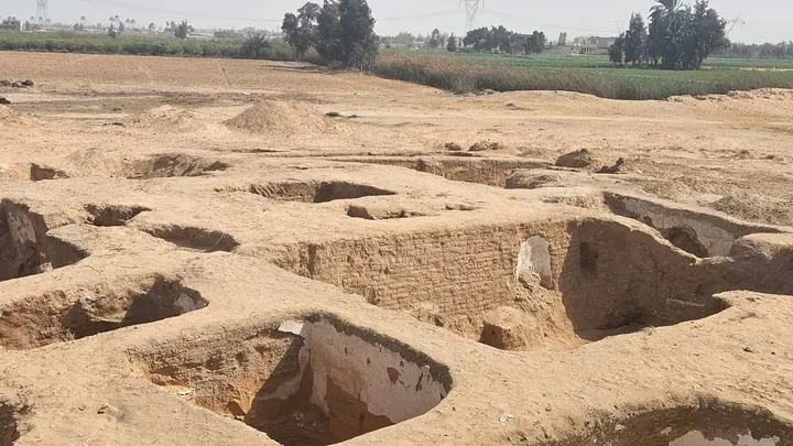 Archaeological site showing excavated foundations of an ancient building with visible mud-brick walls and open pits, surrounded by arid land and distant green fields.