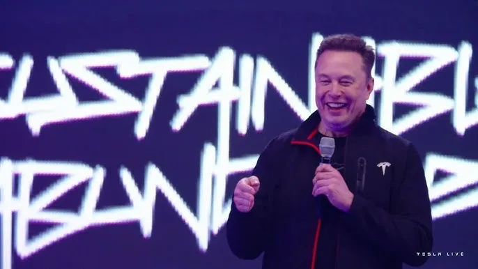 Elon Musk laughing and holding a microphone with a Tesla logo on his jacket.