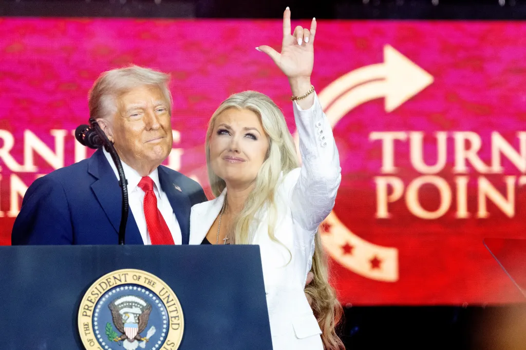 President Trump and Erika Kirk stand together on stage, with Erika making a hand gesture, at the Charlie Kirk Memorial.