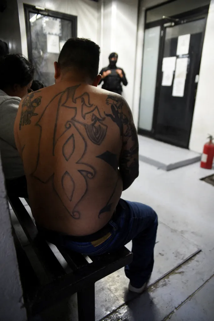 Ever Sinay, a leader of the 18th Street gang, is shown from behind while seated, revealing intricate tattoos covering his back and shoulders, with a police escort visible in the background.