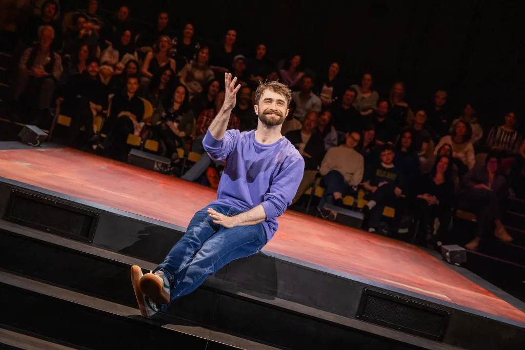 Daniel Radcliffe, with a beard, wearing a purple sweater and jeans, sits on a stage with a red floor, gesturing to a blurry audience.