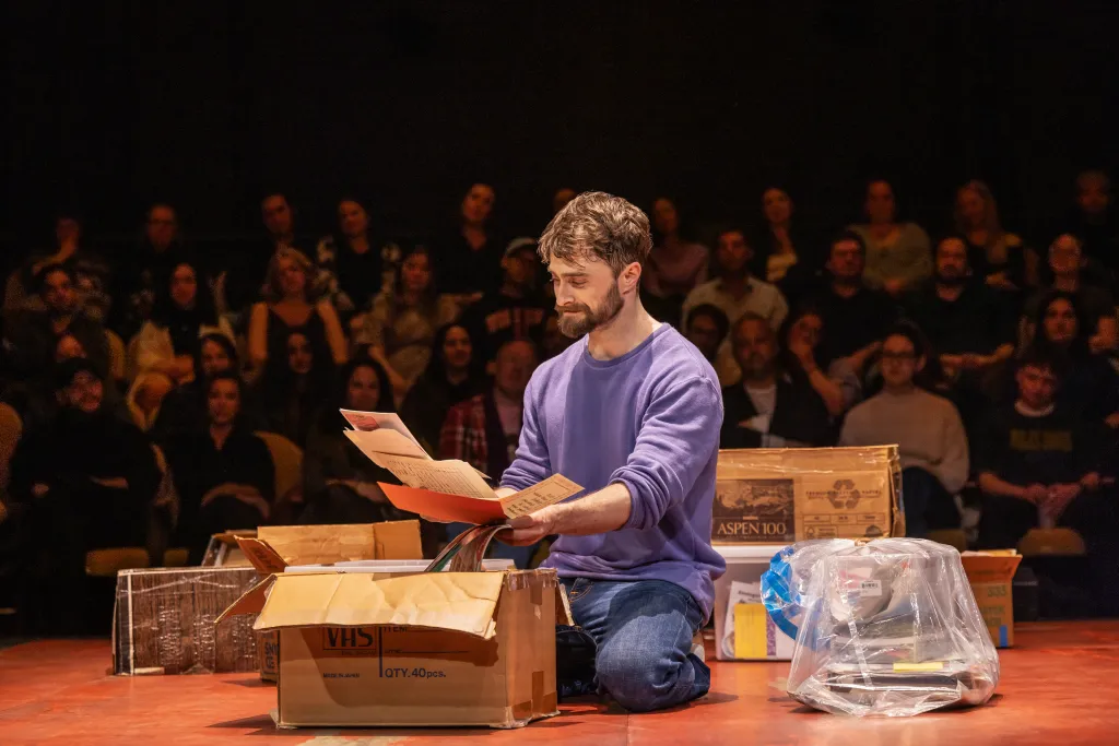 Daniel Radcliffe on his knees, reading notes from a cardboard box, with an audience in the background
