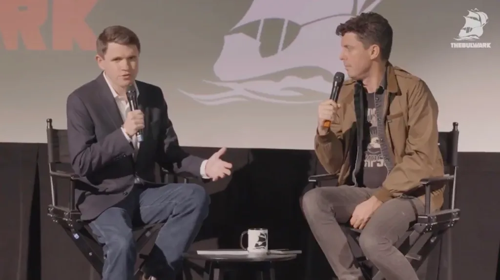 James Talarico and another man speak into microphones while sitting in chairs on a stage.