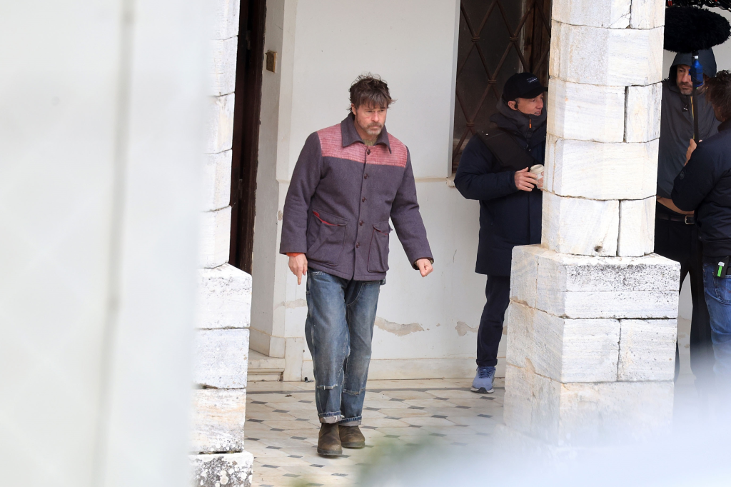Brad Pitt, with tousled hair and a beard, walks in a purple and red jacket on a movie set.