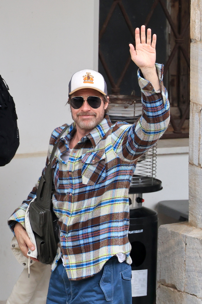 Brad Pitt, dressed in a plaid shirt, sunglasses, and a baseball cap, waves to the camera.