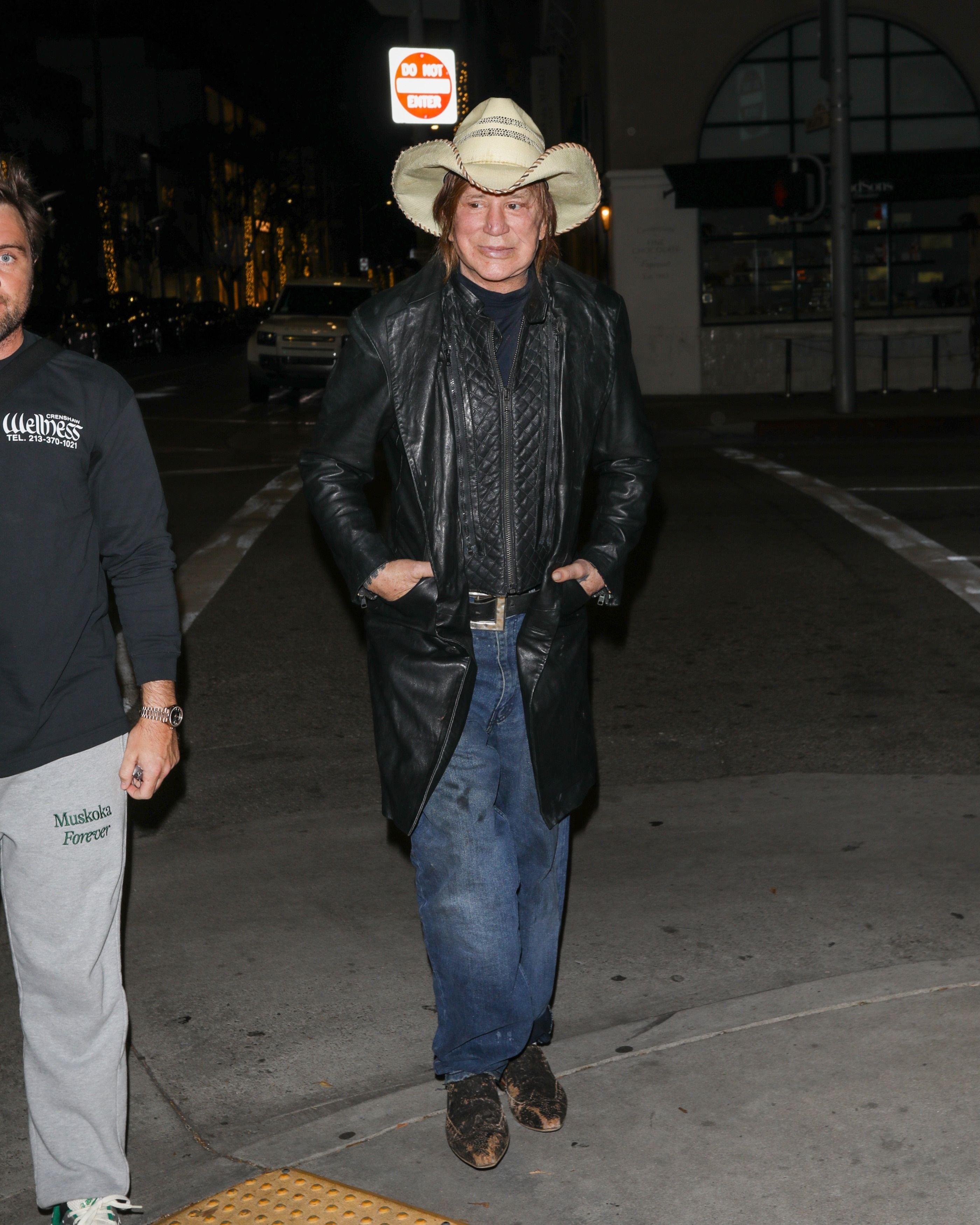 Mickey Rourke walking outdoors at night in a cowboy hat and long black leather coat.