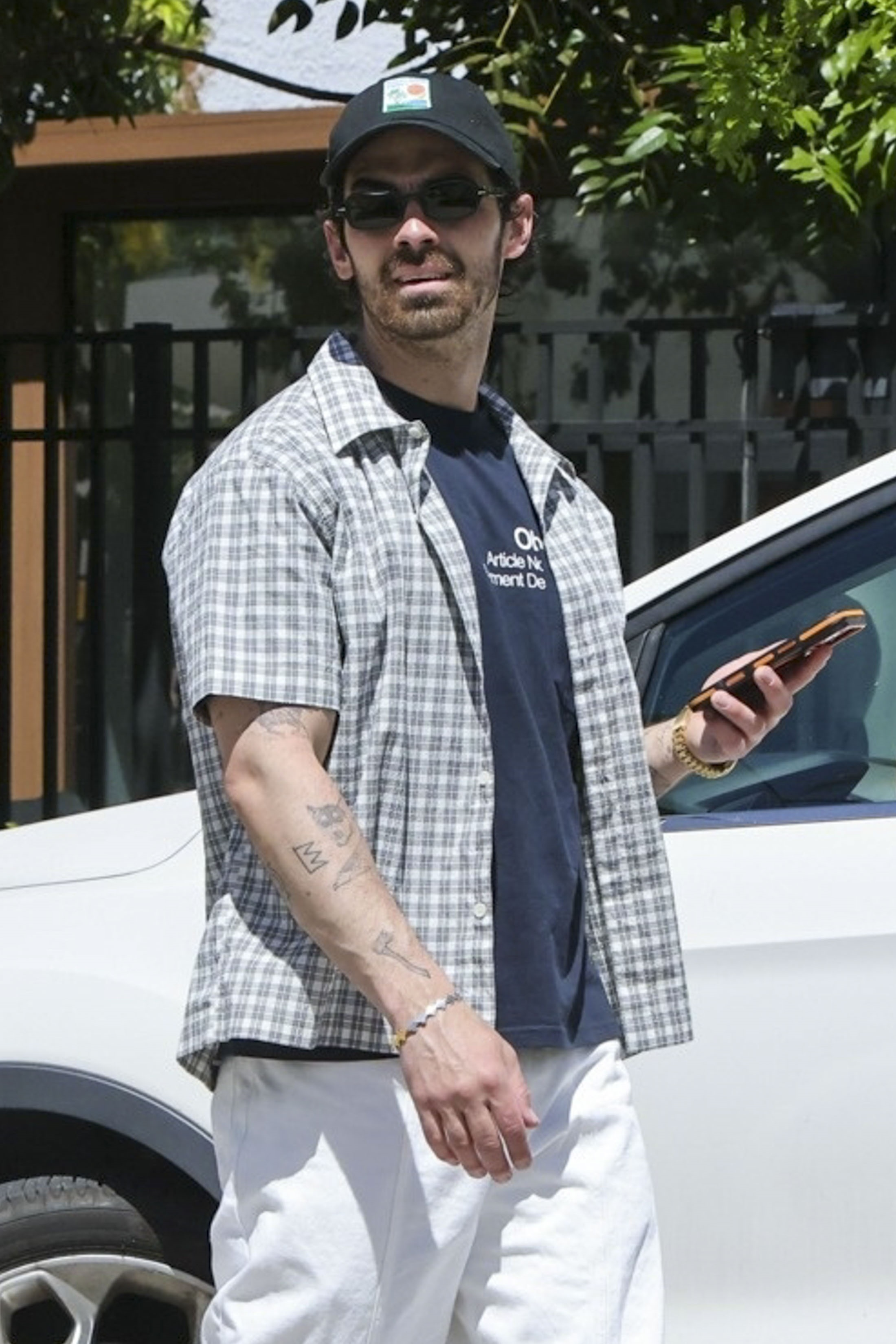 Joe Jonas wearing a plaid shirt, sunglasses, and a baseball cap while holding a phone.