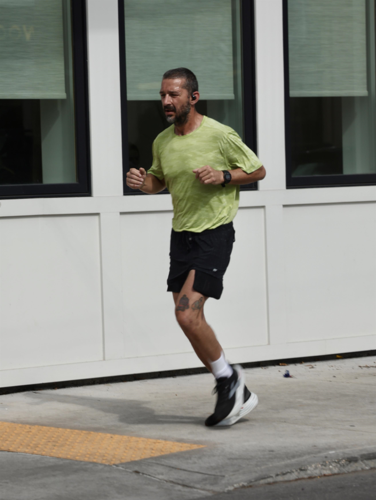 Shia LaBeouf jogging in New Orleans in a light green shirt and black shorts with visible leg tattoos.