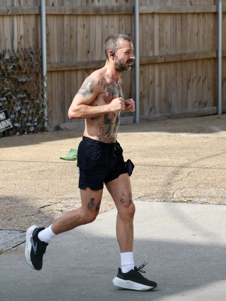 Shia LaBeouf jogging shirtless with numerous tattoos, black shorts, white socks, and black and white athletic shoes.