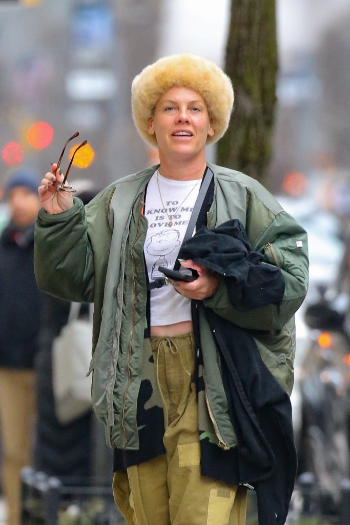Pink walking in NYC in a fur hat, olive jacket, and cargo pants, holding sunglasses and a phone.