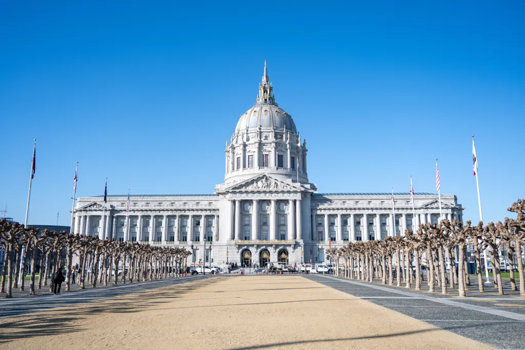 Facade of San Francisco City Hall with a sandy plaza in front and rows of trimmed trees.