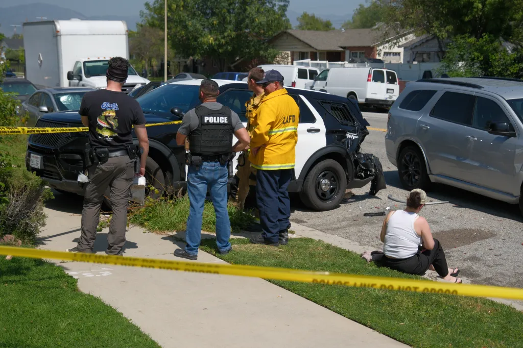 Police officers and a firefighter are on a residential street next to a damaged police SUV and a silver SUV, with yellow police tape in the foreground.