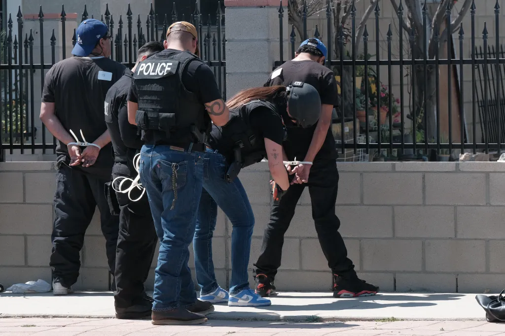 Law enforcement officers arresting individuals in front of a fence.