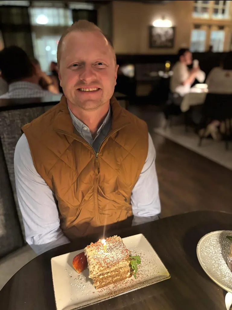 Jason Hughes sits smiling at a table with a birthday cake.