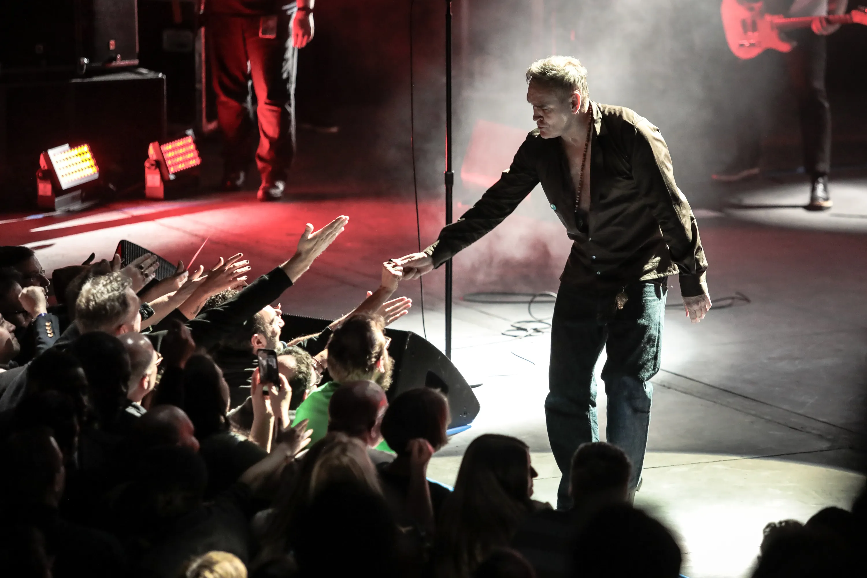 Morrissey reaching his hand into the audience as fans reach back, with stage lighting visible.