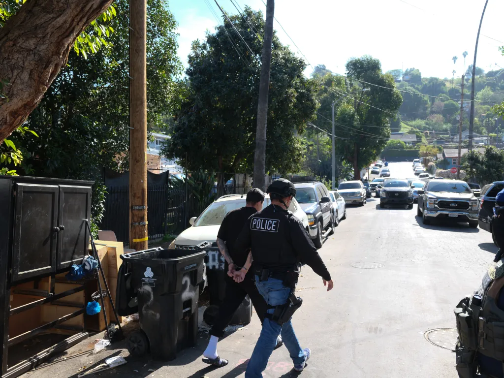 A police officer escorting an arrested, handcuffed man down a street filled with cars.